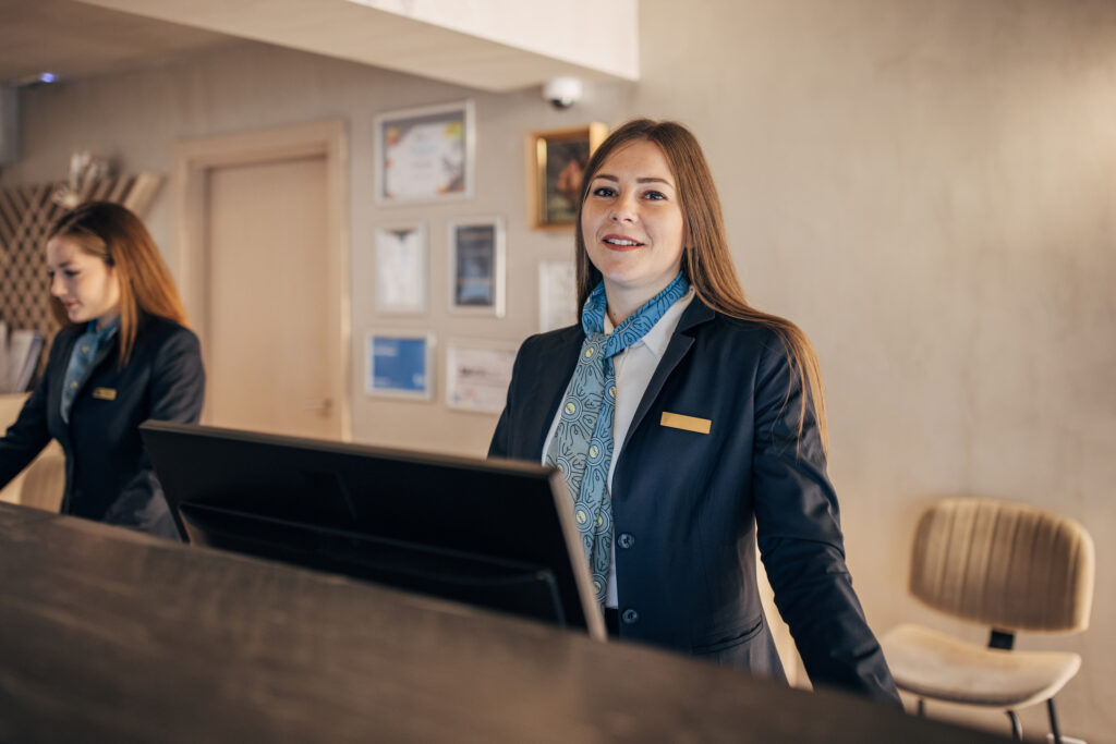 Smiling female hotel receptionist standing in front of a computer monitor.